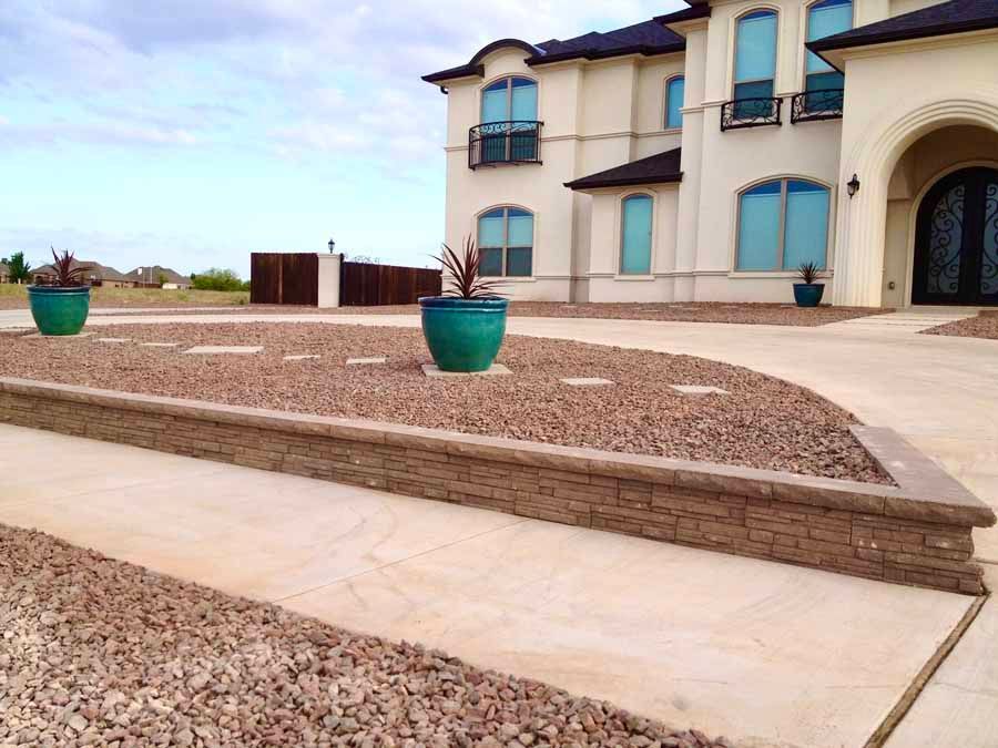 A tan, multi-story house with a rock-filled yard, a low retaining wall, and two teal planters near a concrete walkway.
