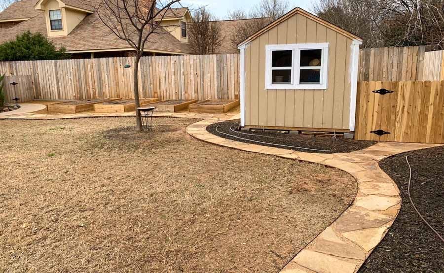 A tan backyard shed sits next to a curved stone path, with raised garden beds and a wooden fence in the background.