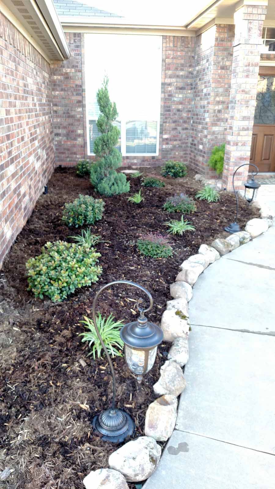 A mulch-covered garden bed beside a brick house, featuring a central evergreen tree, various shrubs, and a stone border.