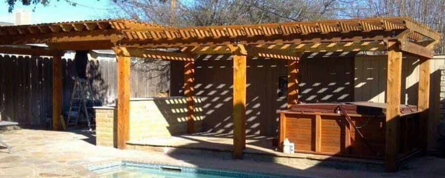 A wooden pergola covers a hot tub and stone outdoor kitchen area by a swimming pool in a backyard setting.