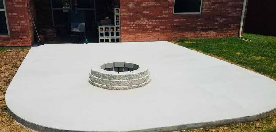 A newly poured concrete patio in a backyard featuring a circular stone fire pit in the center.
