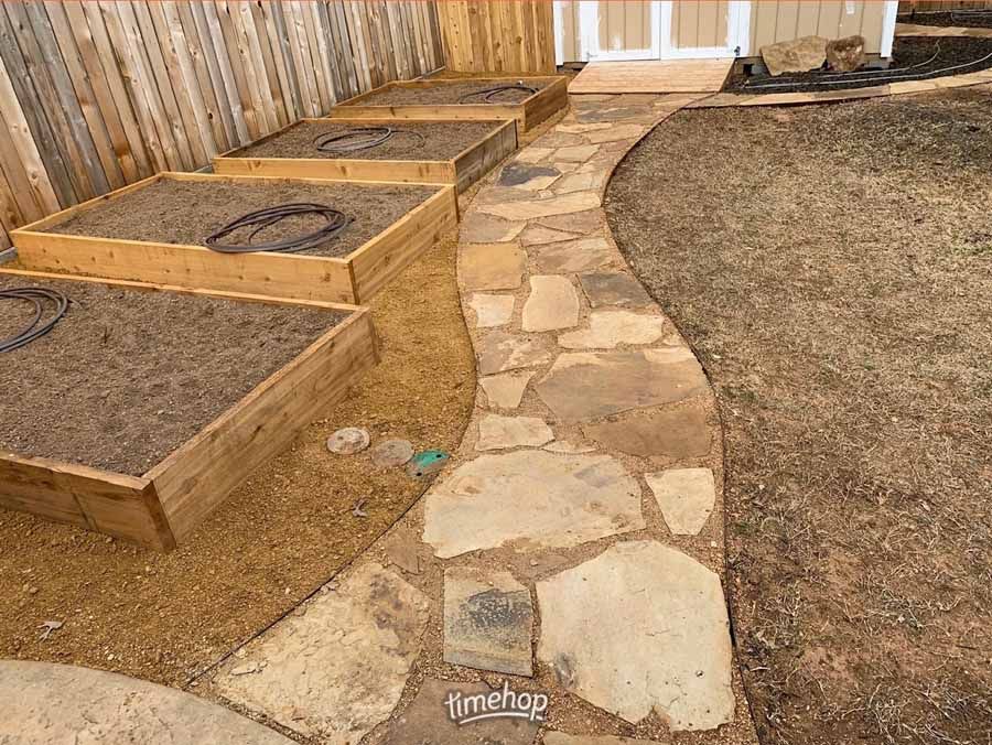 A flagstone path curves past several wooden raised garden beds in a backyard setting.