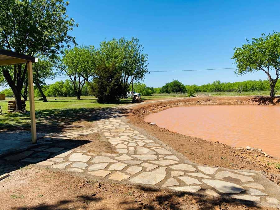 A flagstone walkway curves alongside a muddy pond on a sunny day with green trees in the background.