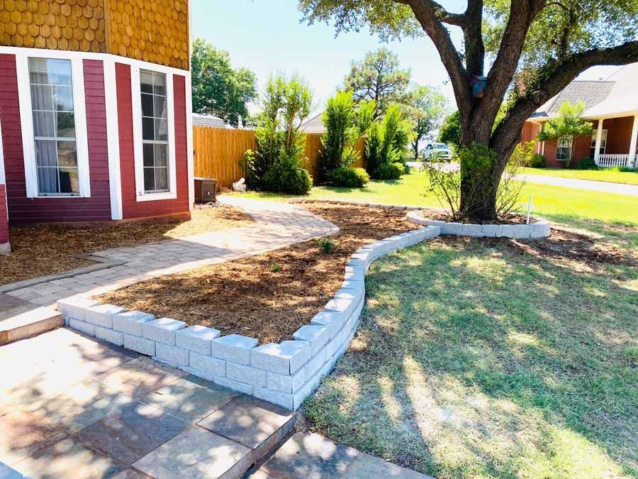 A tiered stone retaining wall border surrounds a mulched garden bed and a large tree in a residential front yard.