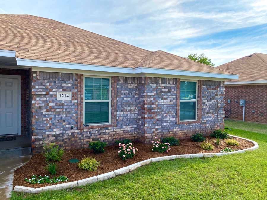 A brick house with a brown roof and a landscaped front garden featuring small shrubs and mulch under a blue sky.