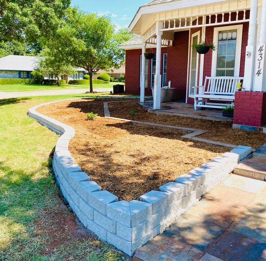 A newly installed gray concrete retaining wall surrounds a mulch-filled garden bed in front of a red brick house.