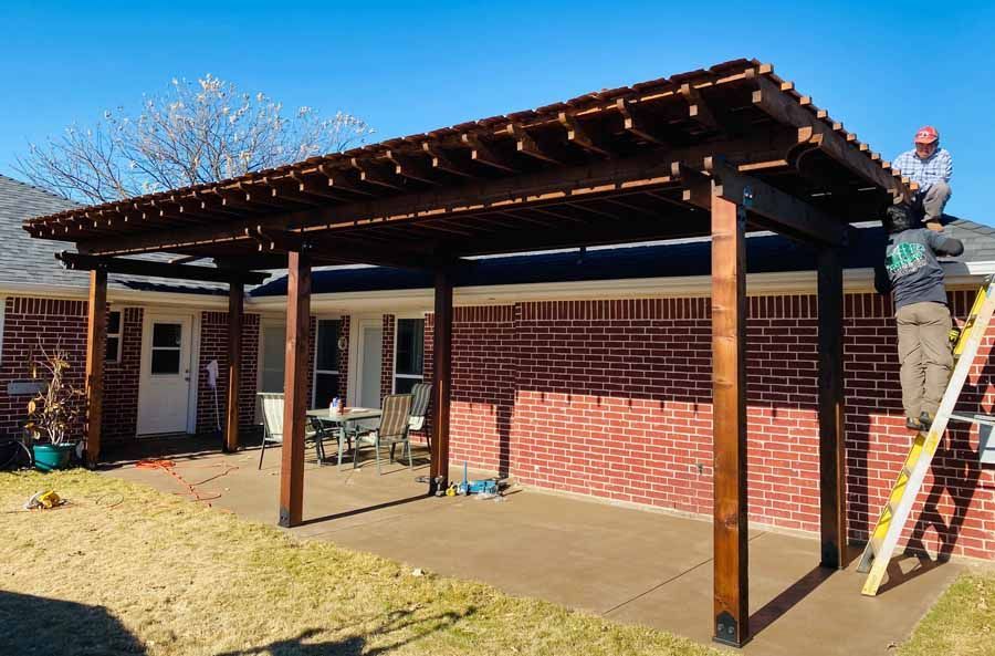 Two workers on a ladder finish installing a dark wood pergola over a residential brick patio.