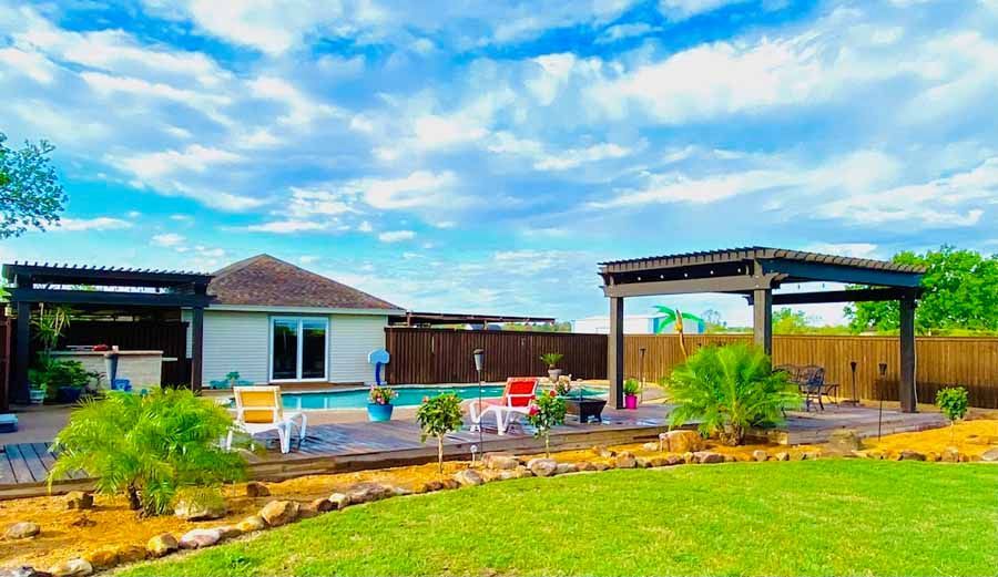 A swimming pool sits in a backyard next to a house, featuring two dark wooden pergolas, lounge chairs, and palm trees.