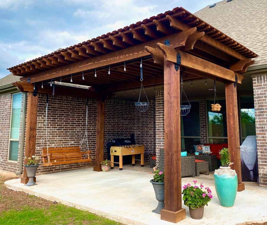 A wooden pergola over a concrete patio featuring a hanging bench swing, a foosball table, and potted plants.
