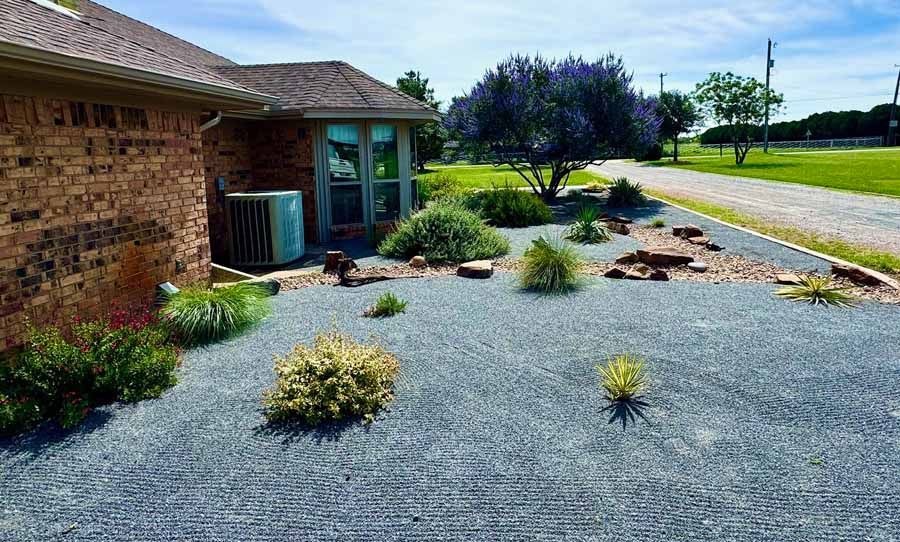 A brick house side exterior with gray gravel landscaping, ornamental grasses, small shrubs, and a blooming purple tree.