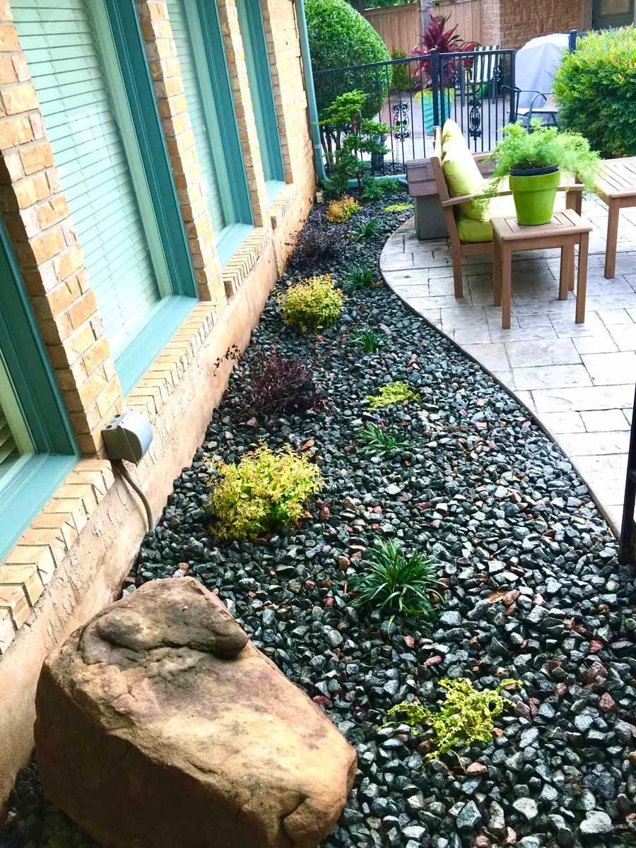 A garden bed with decorative dark gray gravel, small shrubs, and a large boulder along the side of a brick house.