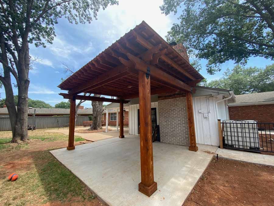A wooden pergola with a slatted roof stands on a concrete patio beside a house with a brick fireplace in a backyard.