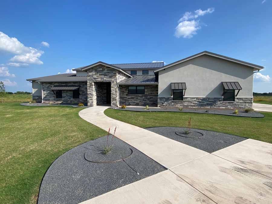 A modern, single-story home with stone siding and light stucco, viewed from a concrete path on a clear, sunny day.