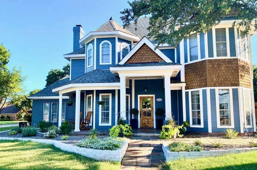 A two-story blue house with a turret, white trim, a covered porch, and brown shingle accents on a sunny day.