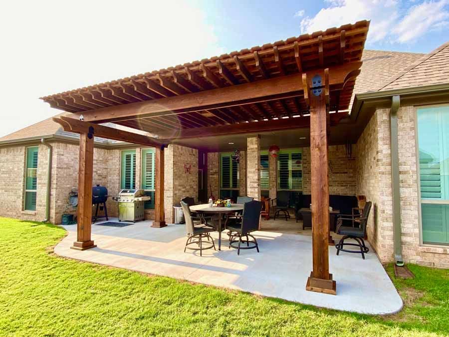 A backyard patio with a wooden pergola, dining set, and grill attached to a brick house.