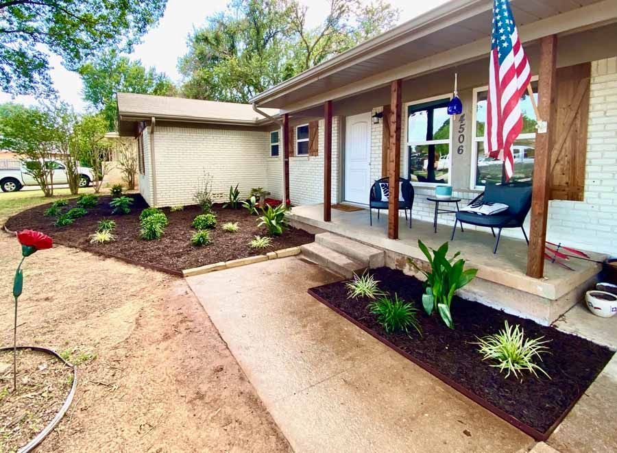 A front view of a white brick house with a concrete walkway, fresh mulch beds, and an American flag on the porch.