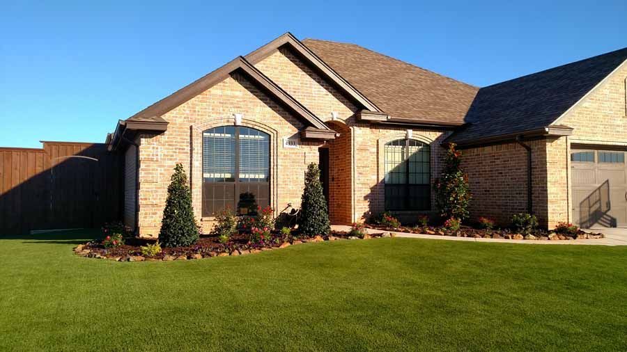 A single-story tan brick house with a brown shingled roof, a manicured lawn, and evergreen bushes under a clear blue sky.