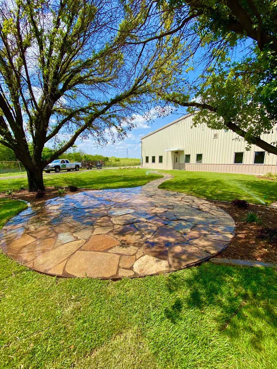 A circular flagstone patio set in a green lawn under two large trees with a light-colored building in the background.