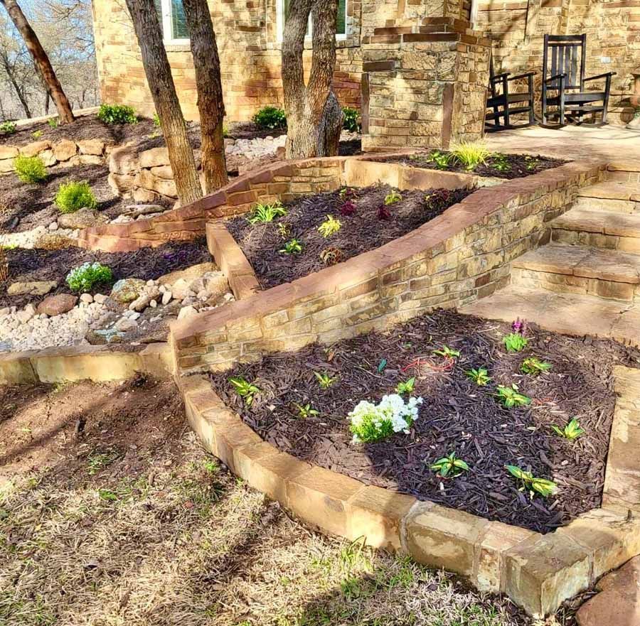 Tiered stone garden beds with mulch and young plants on a stone patio with outdoor chairs near a stone house.