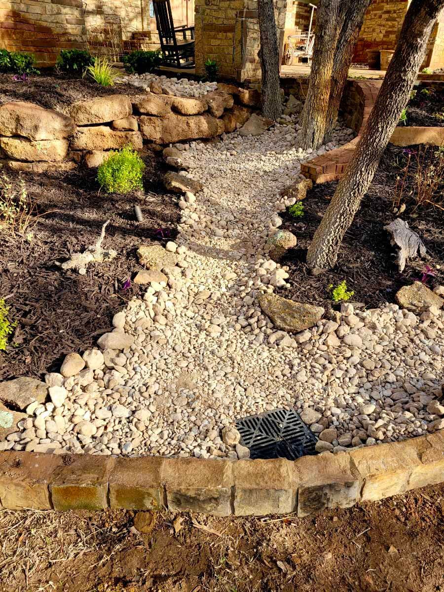 A gravel-lined drainage path with a square drain grate, framed by stones and mulch, leading through a backyard garden.