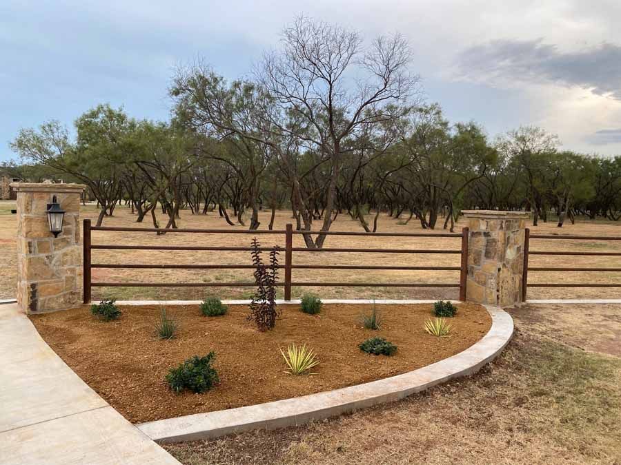 A tan stone garden bed with small desert plants and a metal pipe fence in front of a grove of trees under a cloudy sky.