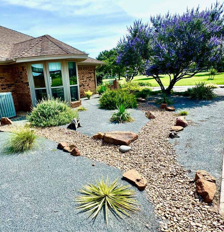 A brick house with a bay window, featuring a xeriscaped front yard with grey gravel, rock accents, and a large purple tree.