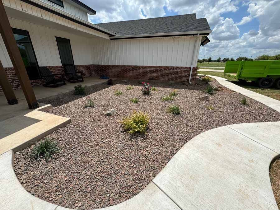 A front yard landscaped with brown gravel and small green shrubs, adjacent to a white house with a brick base.