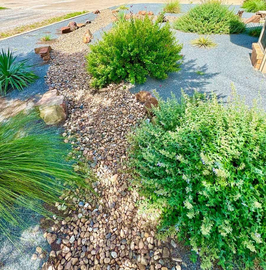 A garden landscaping path of brown river rocks flanked by green bushes and a concrete edge.