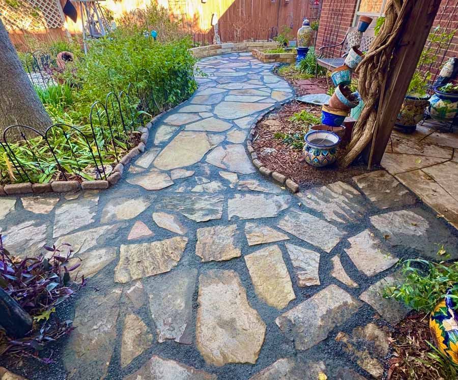 A flagstone path winds through a garden, bordered by brick edging, colorful potted plants, and lush greenery.