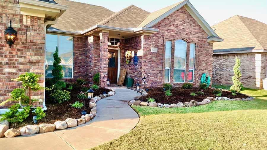 A red-brick house with a curved stone-bordered walkway, mulched flower beds, and green shrubs under a clear blue sky.