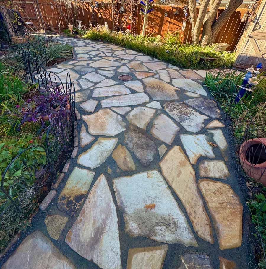 A flagstone path with grey mortar joints winds through a garden, with wooden fencing and plants in the background.