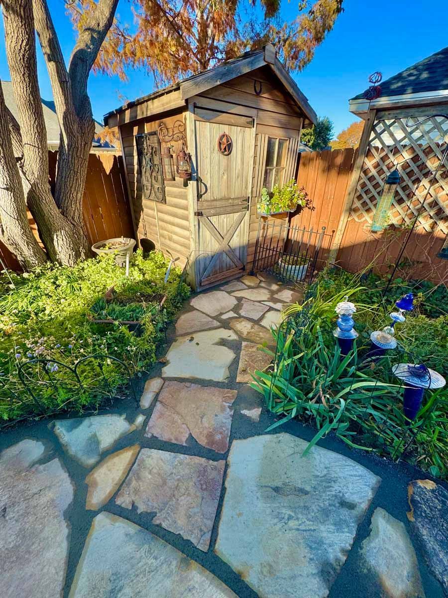 A rustic wooden shed stands at the end of a stone path in a sunlit garden with green foliage and glass garden stakes.