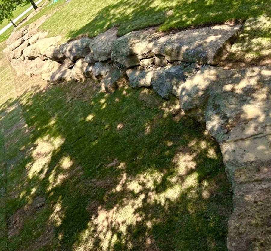 A rough stone retaining wall curves through a grassy lawn, partially shaded by trees.