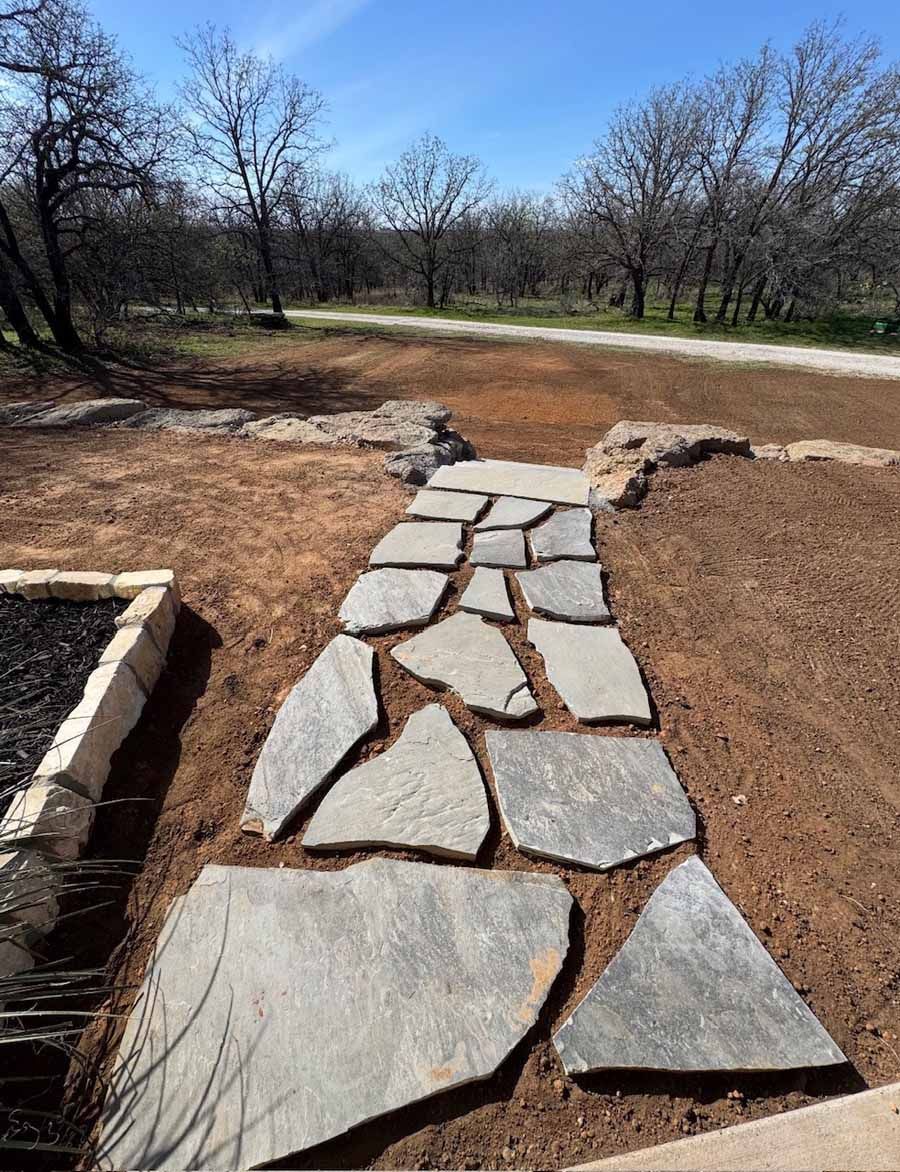 Flagstone pathway leading through a yard toward a gravel path and trees under a clear blue sky.