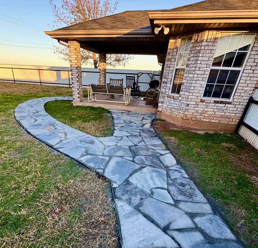 A stone walkway leads to a covered patio with a wooden porch swing next to a brick house at sunset.
