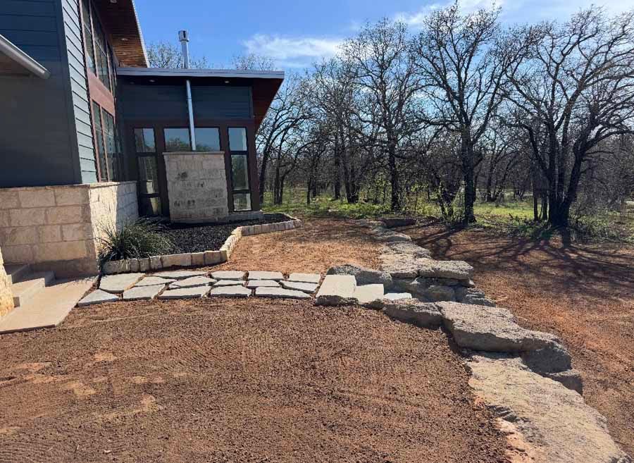 Stone path and retaining wall leading toward a modern home exterior against a background of leafless trees under a blue sky.