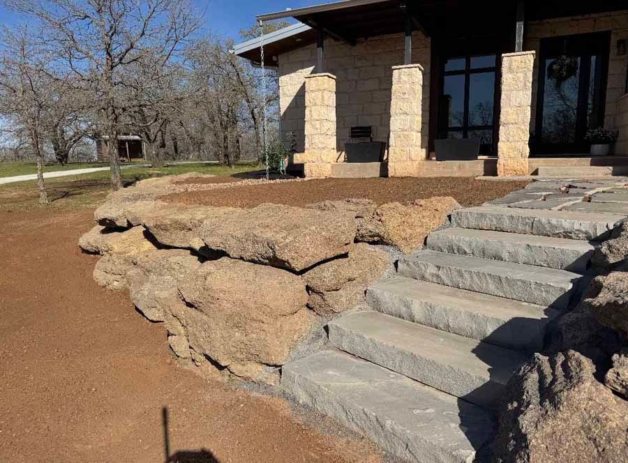 A stone retaining wall with matching gray stone steps leading up to a house entrance on a sunny day.
