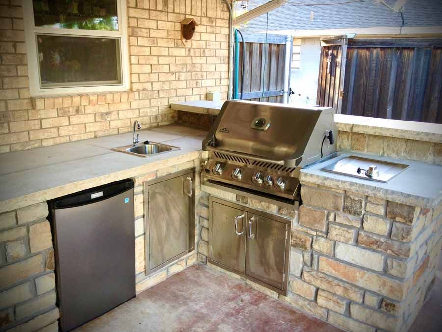 An outdoor kitchen with a stainless steel grill, sink, and mini-fridge set into a stone counter against a brick wall.