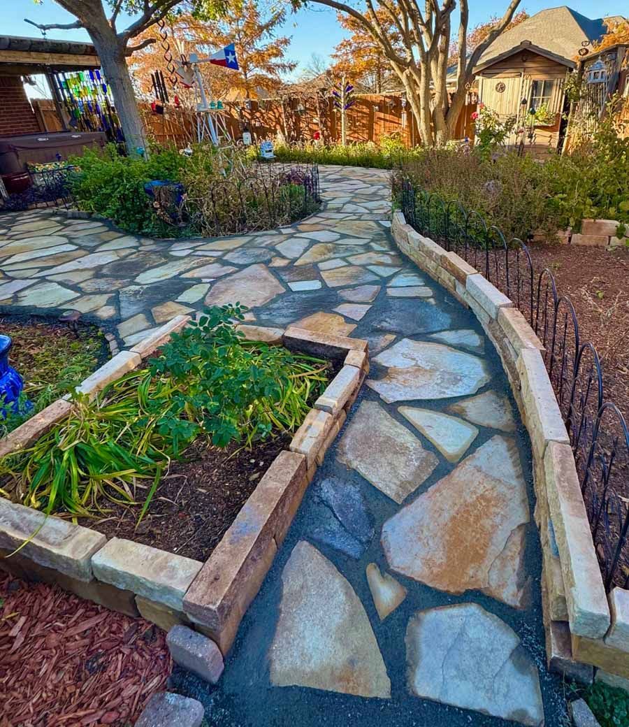 A stone flagstone walkway with a raised garden bed border in a sunny backyard with trees and a house in the background.