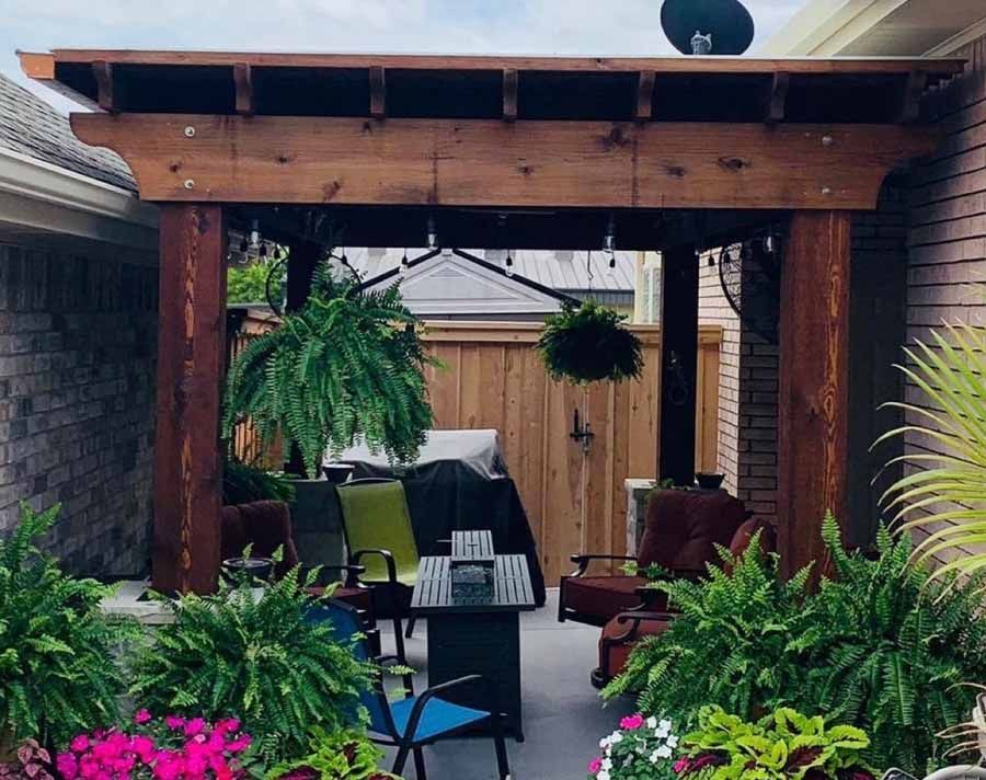 A covered wooden patio pergola with outdoor furniture, lush hanging ferns, and potted plants in a backyard setting.