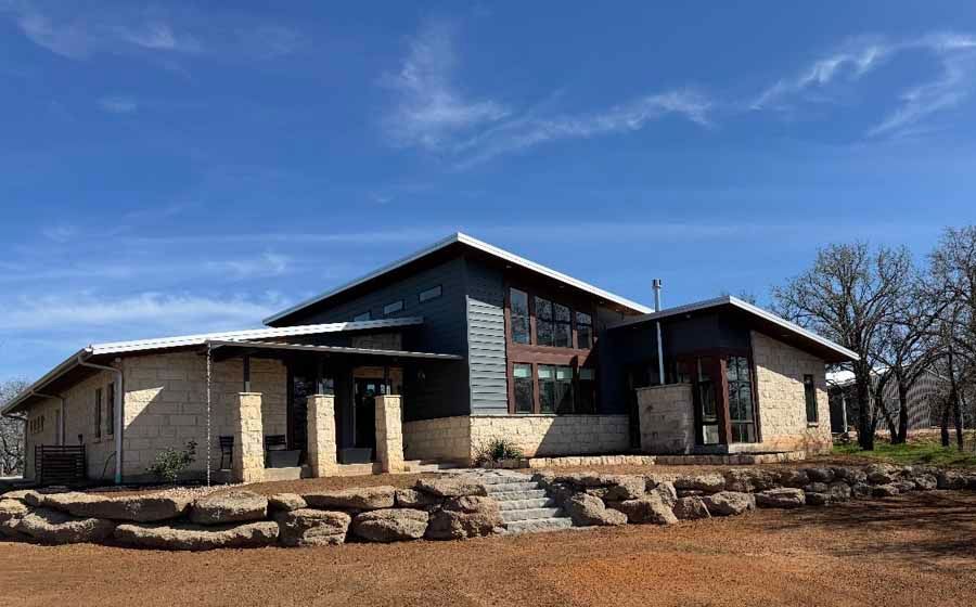 A modern house with stone and dark gray siding, featuring a multi-level roof and stone retaining walls under a blue sky.