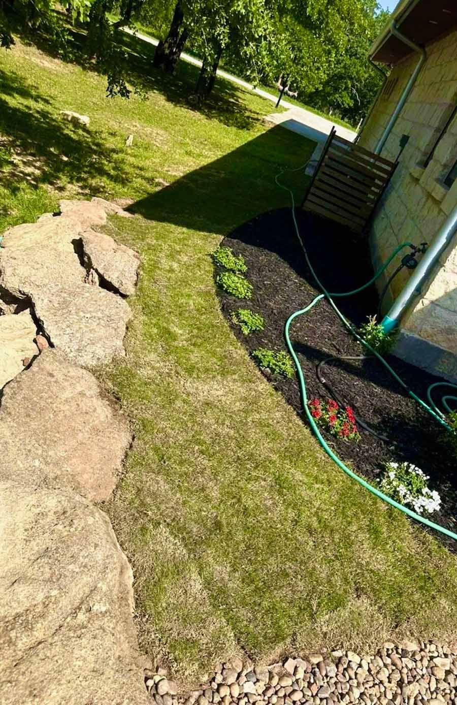 A curved landscaping bed with dark mulch and small plants, bordered by large flat stones and a grassy lawn beside a house.