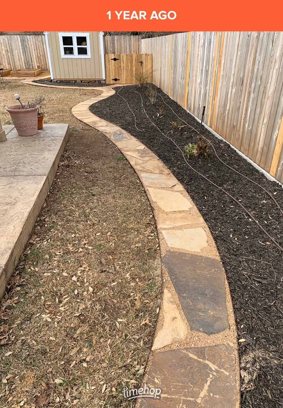 A stone walkway curves through a backyard, bordered by mulch on one side and grass on the other, near a wooden fence.