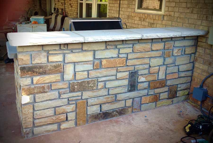 An outdoor bar counter built with tan and brown textured stone veneer, topped with a smooth, light-colored stone slab.