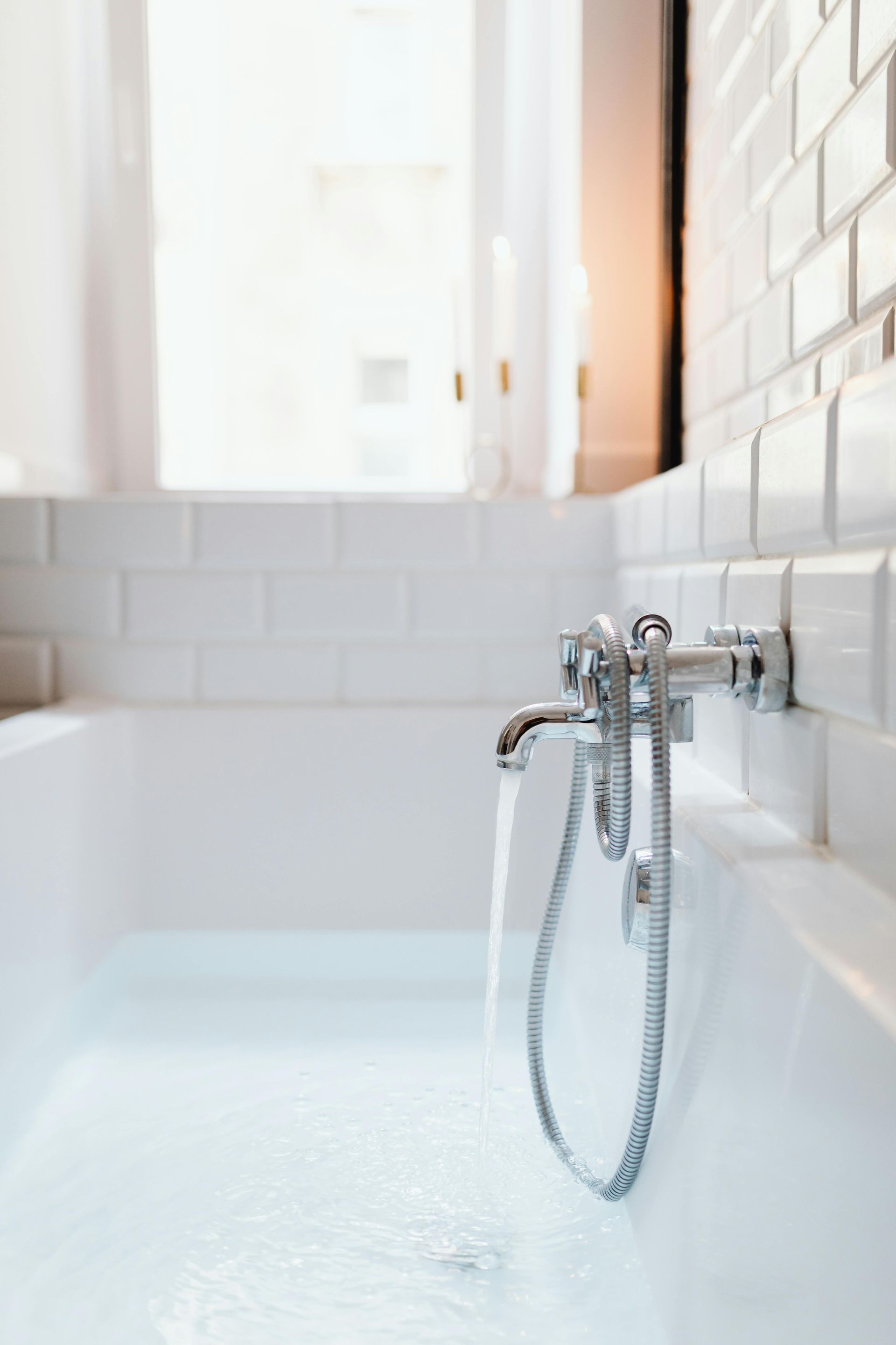 Bathtub filling with water, against white tile wall, near window with sheer curtains.