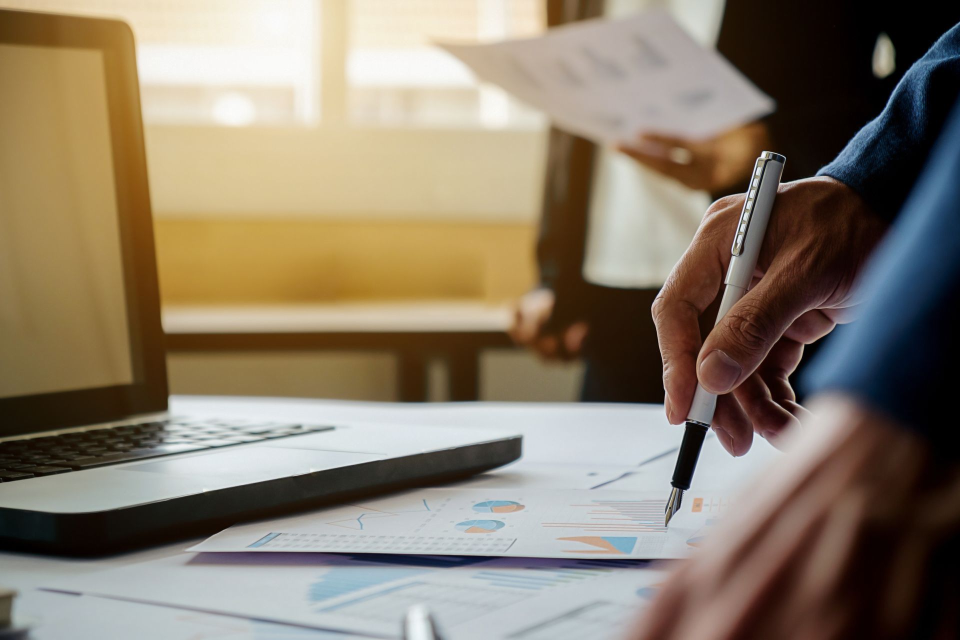 Una persona indica un grafico su un tavolo con un computer portatile; un'altra persona tiene in mano dei fogli sullo sfondo.