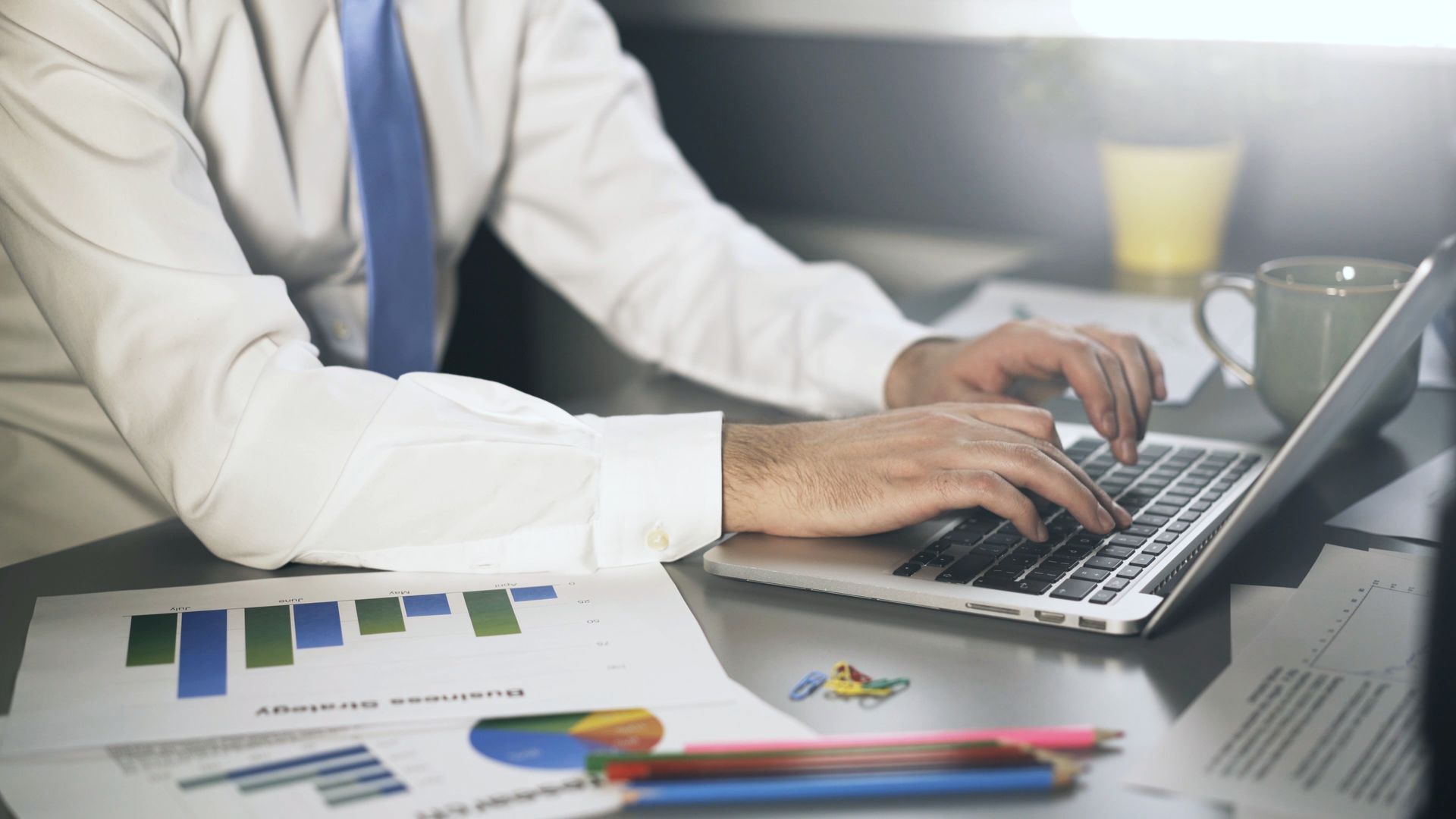 Uomo in camicia bianca e cravatta blu che scrive sul computer portatile alla scrivania con grafici e tazza di caffè.