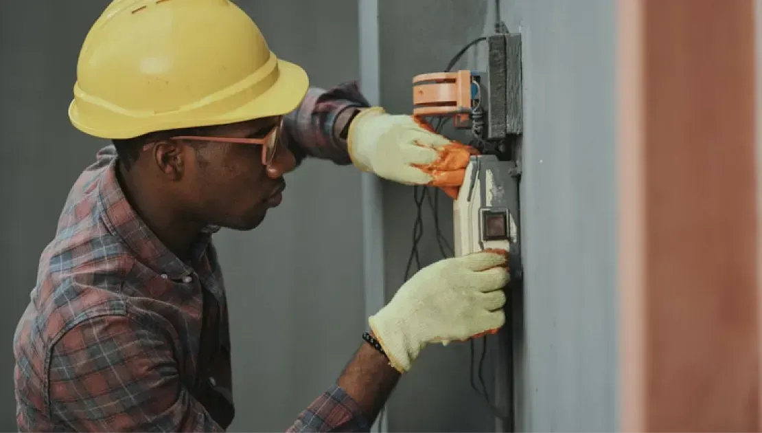 Construction worker in a yellow hard hat and gloves using a tool on a wall-mounted electrical outlet.