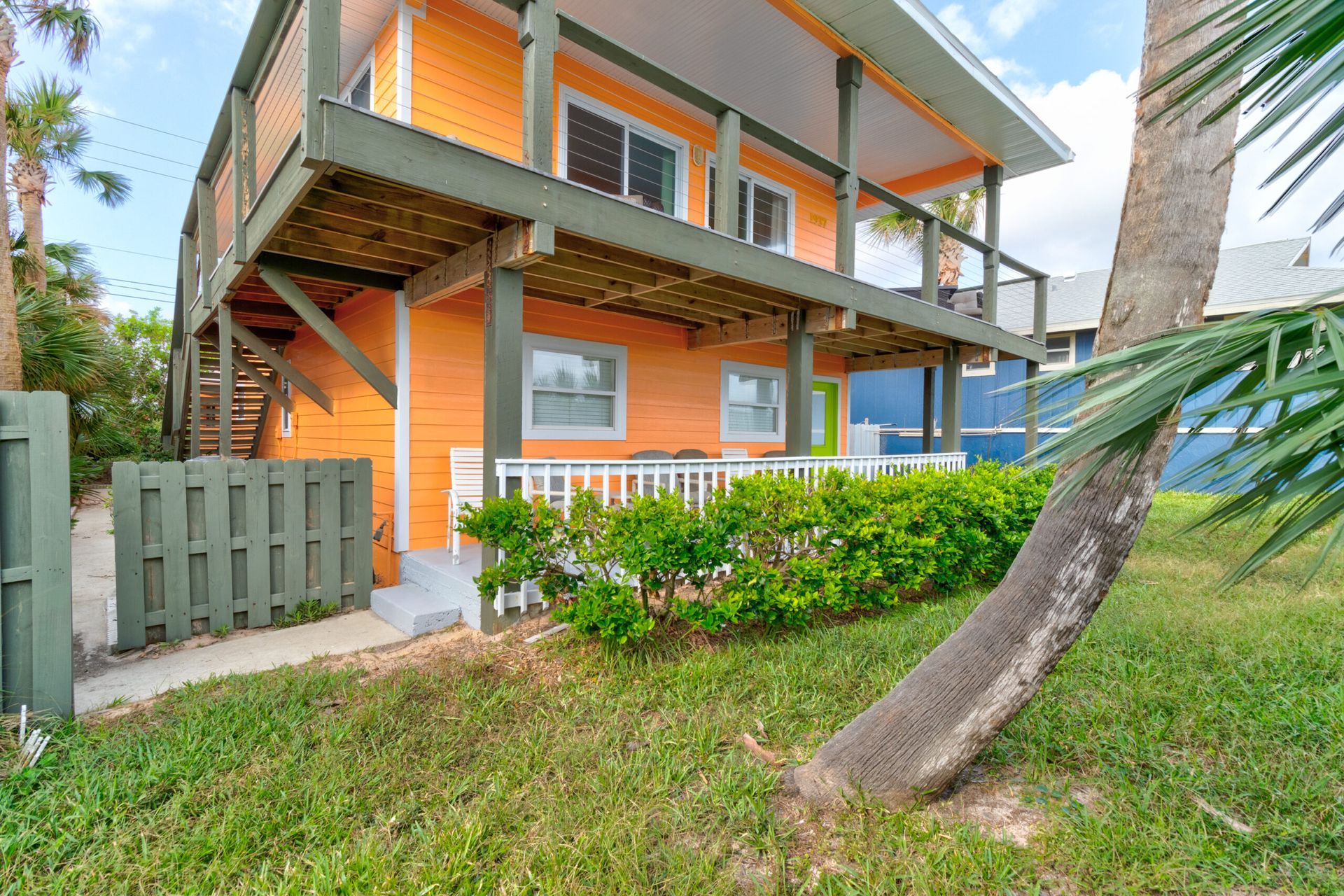 Orange beach house with a second-story deck, green trim, and white railing, near palm trees.