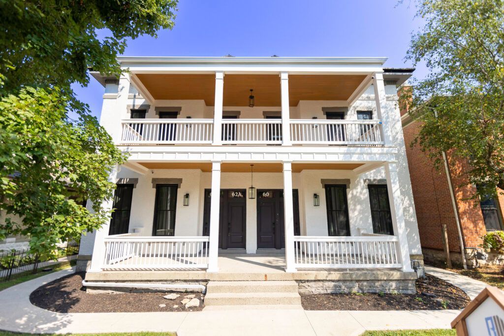 Two-story white building with two front doors and balconies, stone steps, and green lawns.